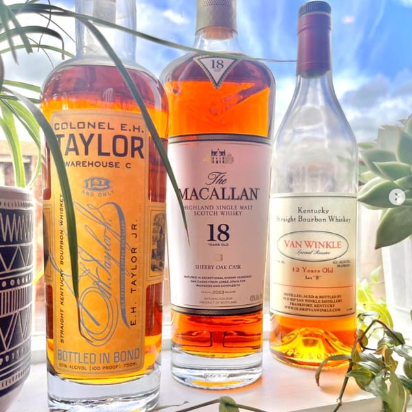 Three bottles of whiskey, including Colonel E.H. Taylor, The Macallan 18, and Van Winkle 12, on a windowsill.
