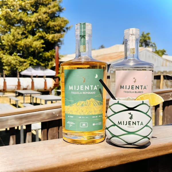 Two Mijenta Tequila bottles and a branded cocktail glass with a lemon wedge on a wooden railing.