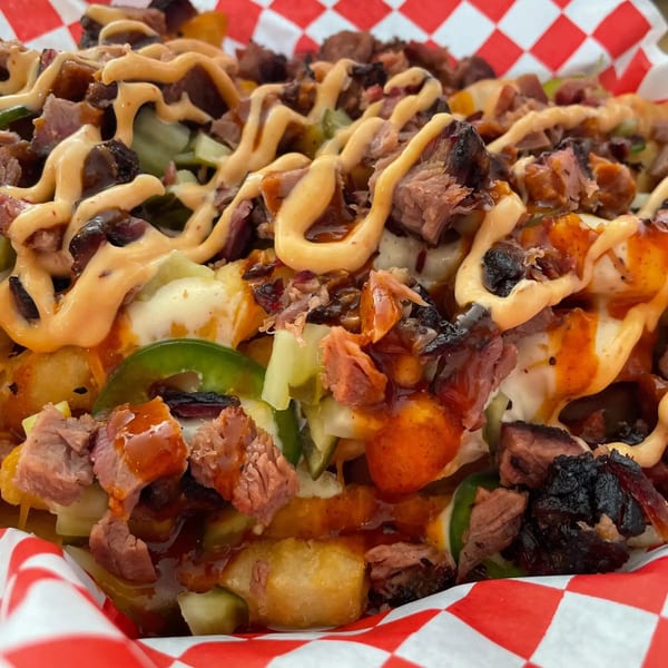 Close-up of BBQ tater tots with pulled pork, cheese sauce, jalapeños, and BBQ sauce in a red checkered basket.